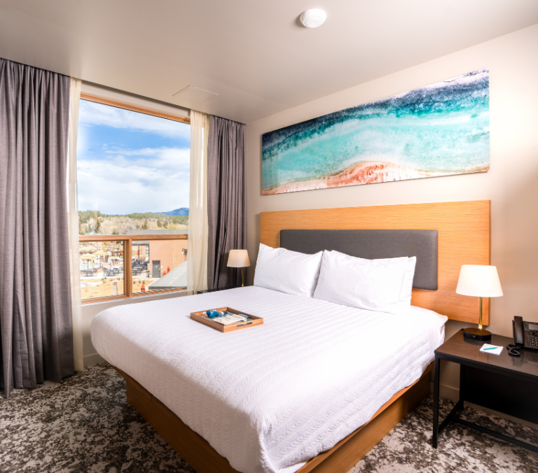 A neatly made bed with a wooden tray holding items sits in a modern hotel room. A large window shows an outdoor view, and an abstract painting hangs above the headboard.