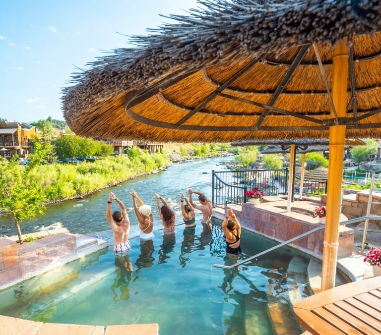 Six people stand in a hot spring pool, arms raised in yoga poses. The pool is next to a river with town buildings and a bridge, under a thatched umbrella.