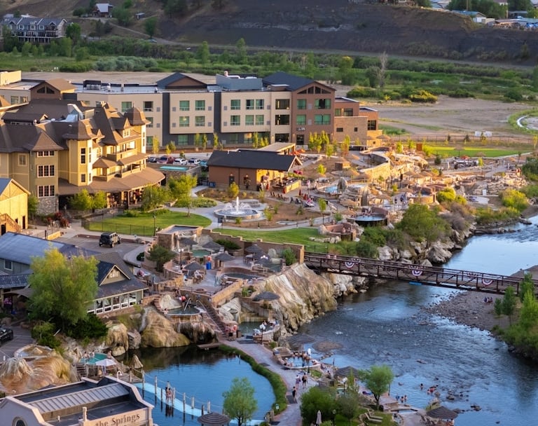 A hot springs resort features numerous pools filled with people alongside a winding river. Nearby are resort buildings, including a 