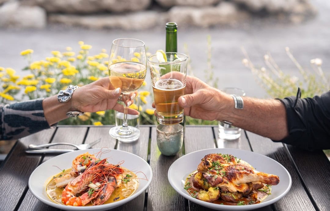 Two people clinking glasses with seafood and chicken dishes at an outdoor resort table.