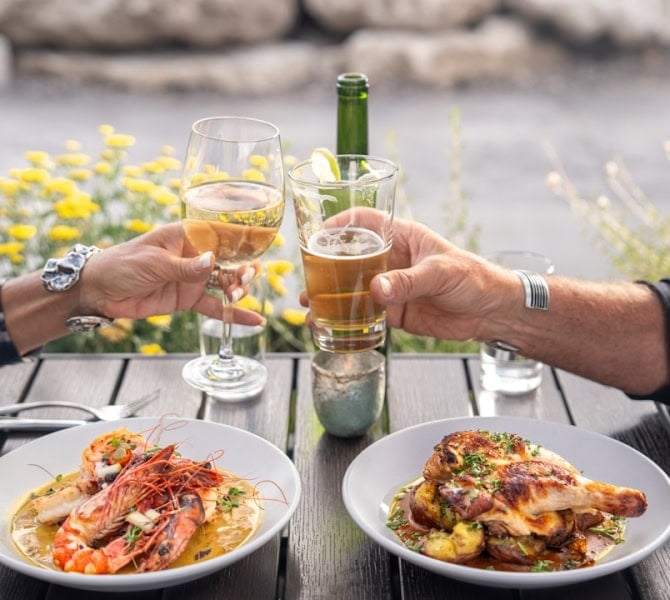 Two people clinking glasses with seafood and chicken dishes at an outdoor resort table.