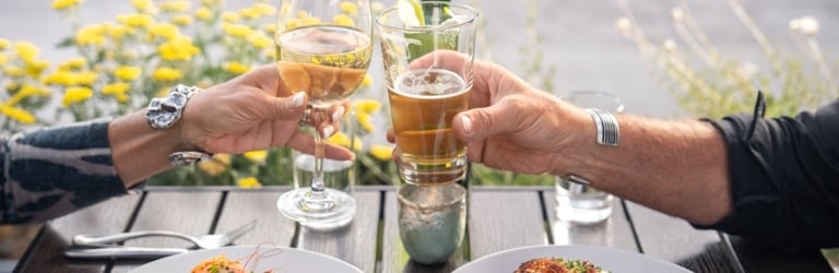 Two hands toast with a white wine glass and a beer glass over shrimp and chicken dishes on a dark outdoor table, with yellow flowers behind.