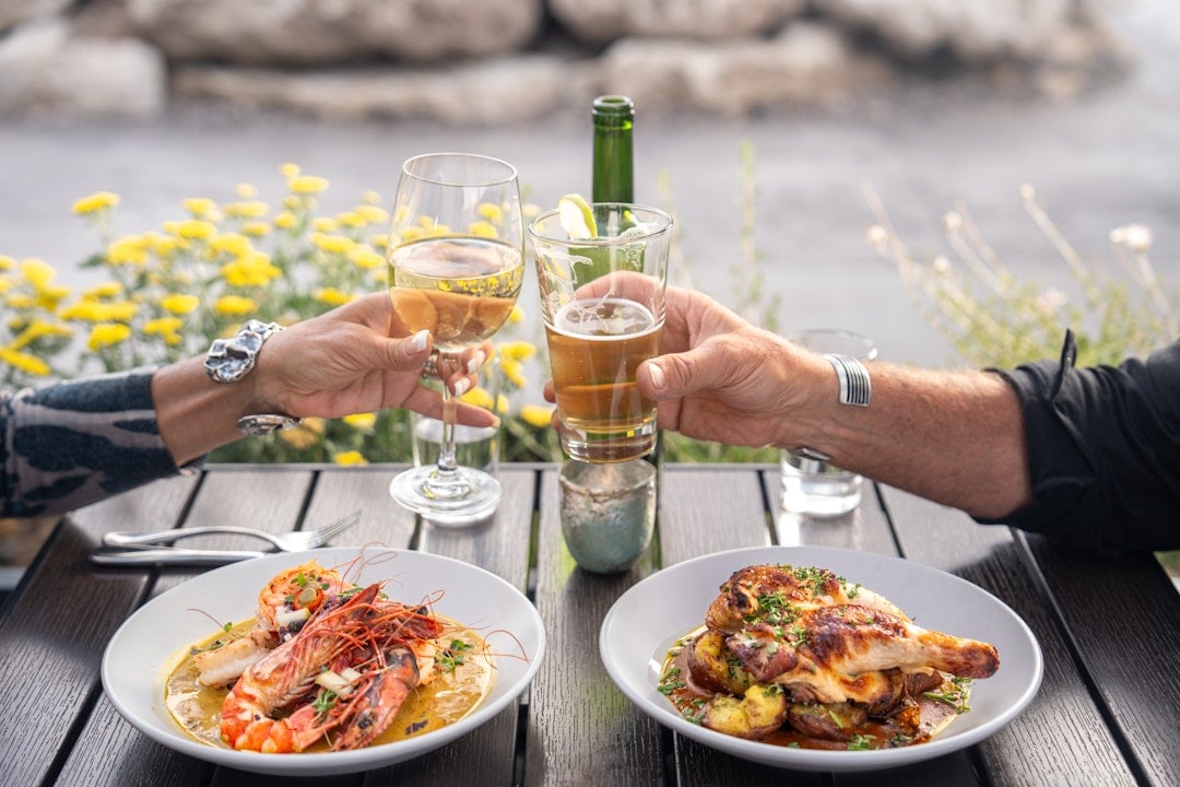 Two hands toast with a white wine glass and a beer glass over shrimp and chicken dishes on a dark outdoor table, with yellow flowers behind.