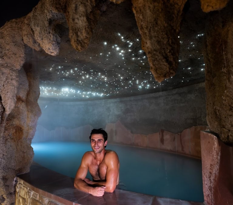 A man smiles, leaning on the edge of a light blue pool, surrounded by dark, cave-like rock formations with a glowing, star-like ceiling.