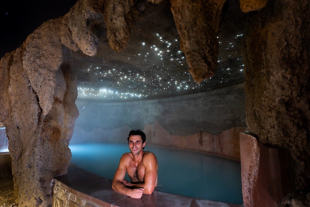 A man smiles, leaning on the edge of a light blue pool, surrounded by dark, cave-like rock formations with a glowing, star-like ceiling.
