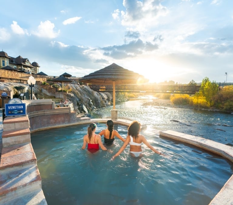 Three women relax in a hot spring pool beside a river, with a bridge, building complex, and autumn foliage under a sunset. A sign reads: 