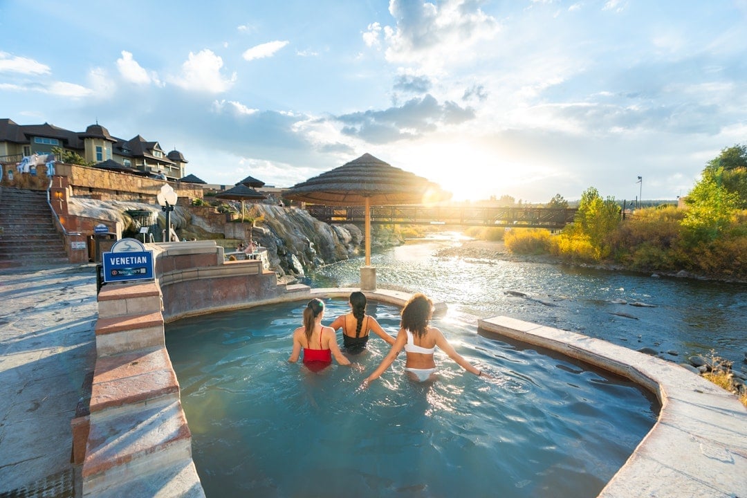 Three women relax in a hot spring pool beside a river, with a bridge, building complex, and autumn foliage under a sunset. A sign reads: 