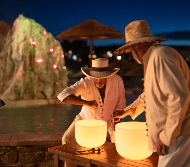 Two men in hats play glowing crystal singing bowls outdoors at night. They stand by a pool with an illuminated rock feature and resort umbrellas.