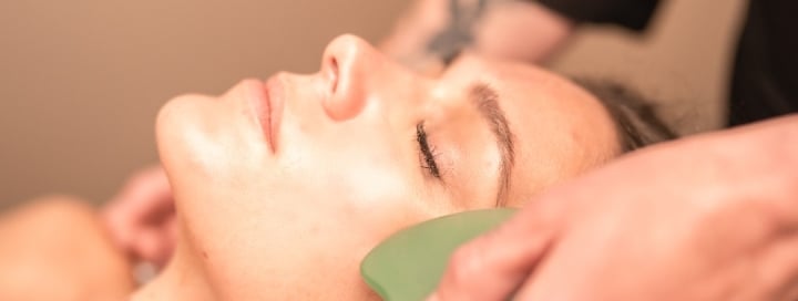 A woman's face, eyes closed, receives a facial. A hand glides a green Gua Sha tool along her cheek/jawline as she lies relaxed during a treatment.