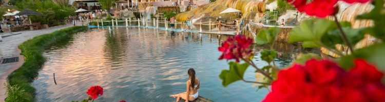 A woman in a white bikini sits on a wooden dock, dipping her feet into a large pool. Behind her are hot springs, a pink hotel, and mountains. Red flowers frame the foreground.
