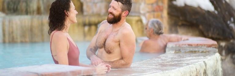 A smiling man and woman chat in a light-blue hot spring pool. Steam rises as rock walls and snow-covered cliffs frame the background.