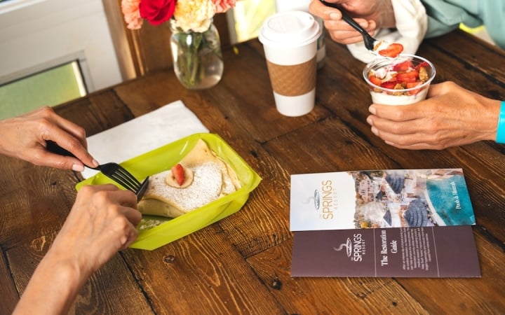 Two people eat breakfast on a wooden table: one cuts a crepe with a strawberry, another spoons yogurt parfait, alongside a coffee cup and flowers.

Text:
THE SPRINGS RESORT
Pools & Minerals
THE SPRINGS RESORT
The Restoration Guide
After an intense day of activity, kick-start your body's natural recovery process. This restoration journey will allow you to maximize your time while visiting The Springs Resort and Spa. Our unique blend of natural mineral water will help flush toxins and muscular micro-tears from intense exercise.
