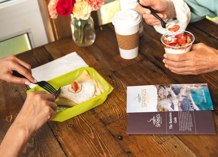 Two people eat breakfast on a wooden table: one cuts a crepe with a strawberry, another spoons yogurt parfait, alongside a coffee cup and flowers.

Text:
THE SPRINGS RESORT
Pools & Minerals
THE SPRINGS RESORT
The Restoration Guide
After an intense day of activity, kick-start your body's natural recovery process. This restoration journey will allow you to maximize your time while visiting The Springs Resort and Spa. Our unique blend of natural mineral water will help flush toxins and muscular micro-tears from intense exercise.