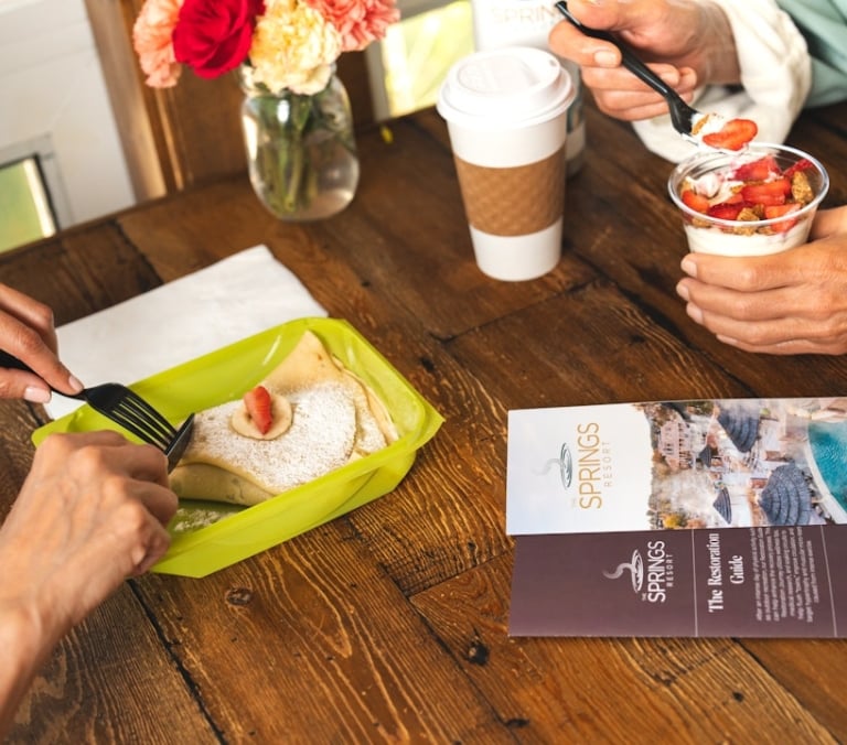 Two people eat breakfast on a wooden table: one cuts a crepe with a strawberry, another spoons yogurt parfait, alongside a coffee cup and flowers.

Text:
THE SPRINGS RESORT
Pools & Minerals
THE SPRINGS RESORT
The Restoration Guide
After an intense day of activity, kick-start your body's natural recovery process. This restoration journey will allow you to maximize your time while visiting The Springs Resort and Spa. Our unique blend of natural mineral water will help flush toxins and muscular micro-tears from intense exercise.
