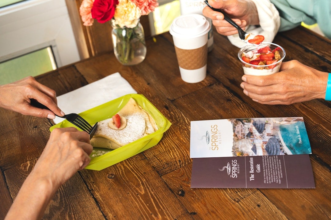 Two people eat breakfast on a wooden table: one cuts a crepe with a strawberry, another spoons yogurt parfait, alongside a coffee cup and flowers.

Text:
THE SPRINGS RESORT
Pools & Minerals
THE SPRINGS RESORT
The Restoration Guide
After an intense day of activity, kick-start your body's natural recovery process. This restoration journey will allow you to maximize your time while visiting The Springs Resort and Spa. Our unique blend of natural mineral water will help flush toxins and muscular micro-tears from intense exercise.