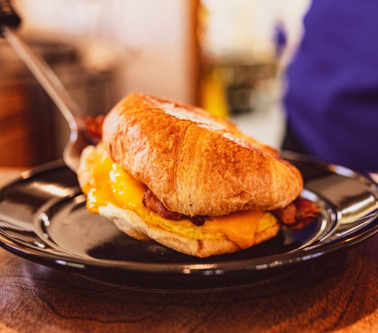 A croissant breakfast sandwich with egg, melted cheese, and bacon is being placed onto a black plate with a metal spatula, on a wooden surface.