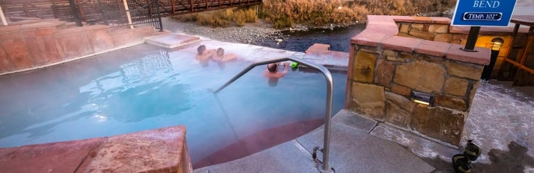 People soak in a steaming hot spring pool next to a river with a town and bridge in the background. A sign reads: RIVER BEND, TEMP: 102°.