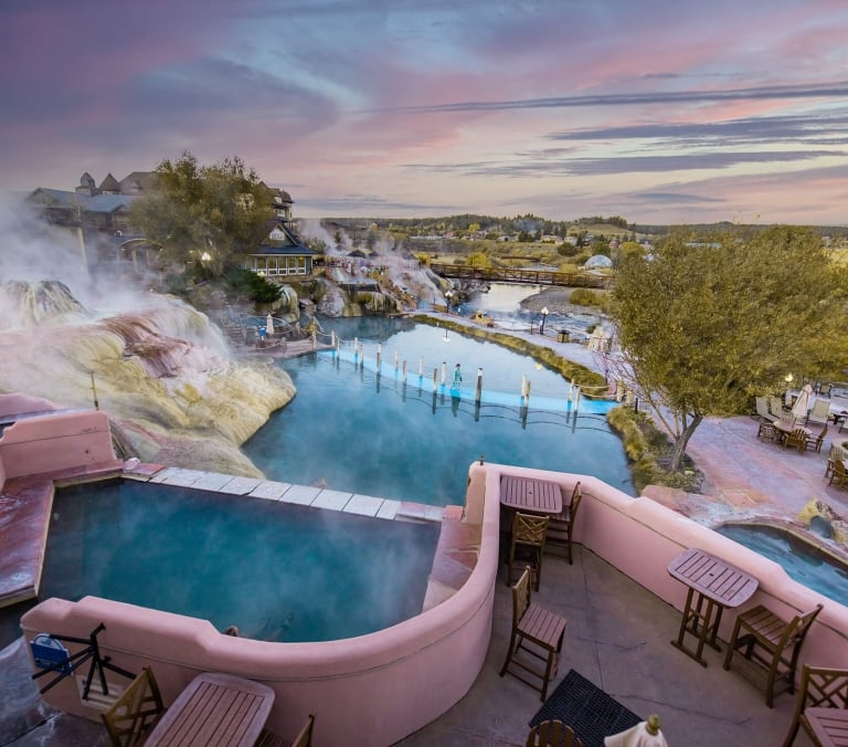 Natural hot springs and multiple turquoise pools steam. Water flows between unique rock formations, surrounded by resort buildings, a river, and walkways under a sunset sky.