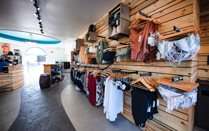 Beachwear hangs on wooden walls and shelves inside a bright retail store with a curved counter and an archway exit. Text: EXIT.