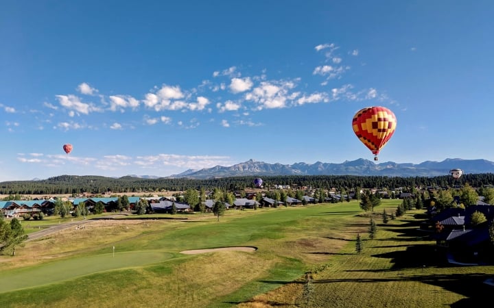 Hot air balloons float above a sprawling green golf course and residential area, set against a backdrop of distant mountains under a bright blue sky.