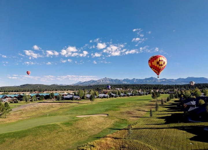 Hot air balloons float above a sprawling green golf course and residential area, set against a backdrop of distant mountains under a bright blue sky.