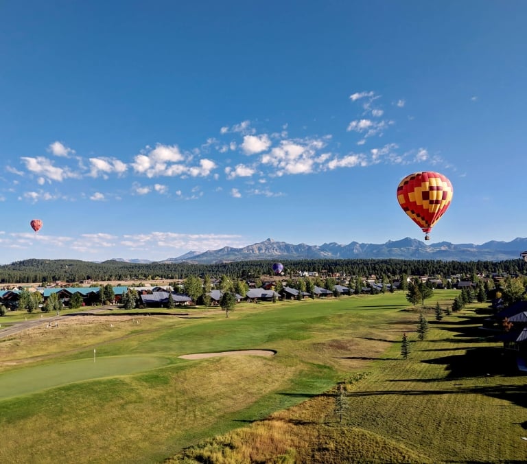 Hot air balloons float above a sprawling green golf course and residential area, set against a backdrop of distant mountains under a bright blue sky.