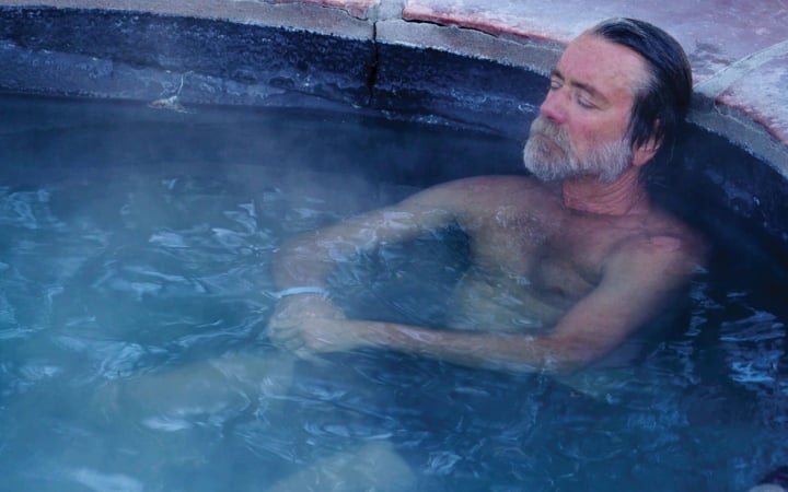 A bearded man relaxes with closed eyes in a steaming, blue-tinted hot tub with rough, reddish-brown stone edges.