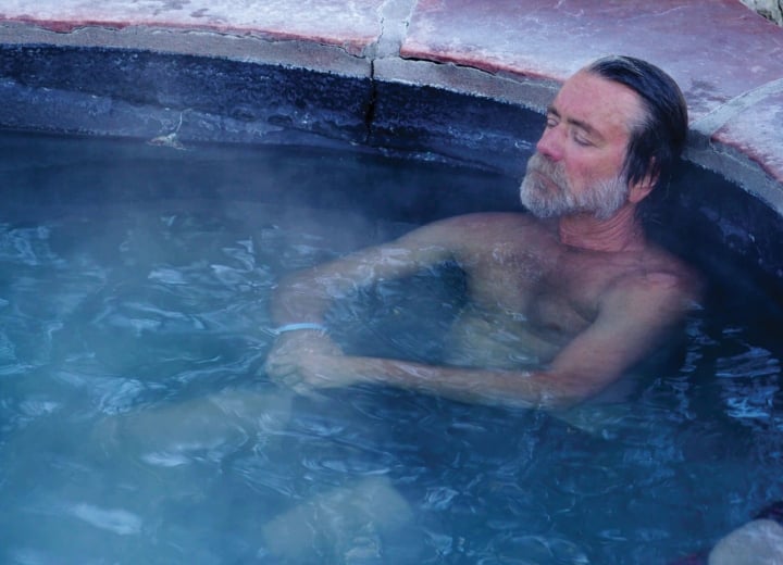A bearded man relaxes with closed eyes in a steaming, blue-tinted hot tub with rough, reddish-brown stone edges.