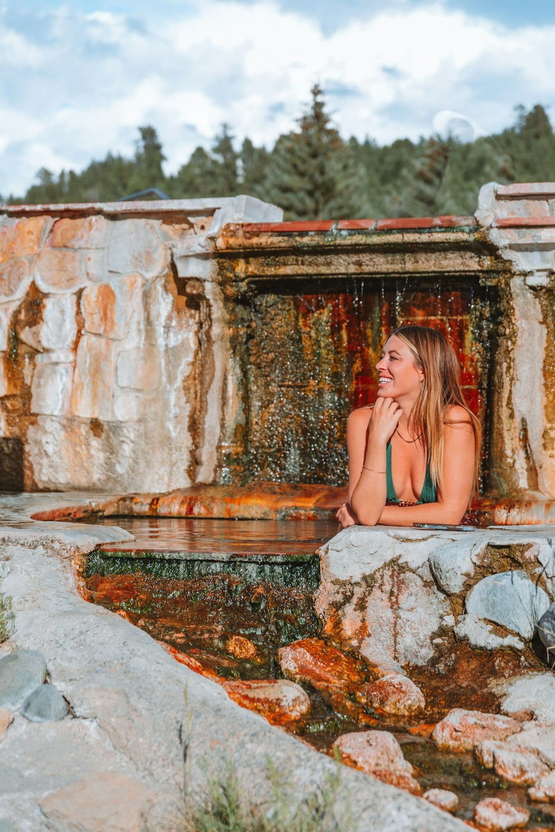 A smiling woman relaxes in a mineral-rich hot spring pool, leaning on the stone edge. Water cascades from a textured rock wall behind her, set amidst rocks and pine trees under a cloudy sky.