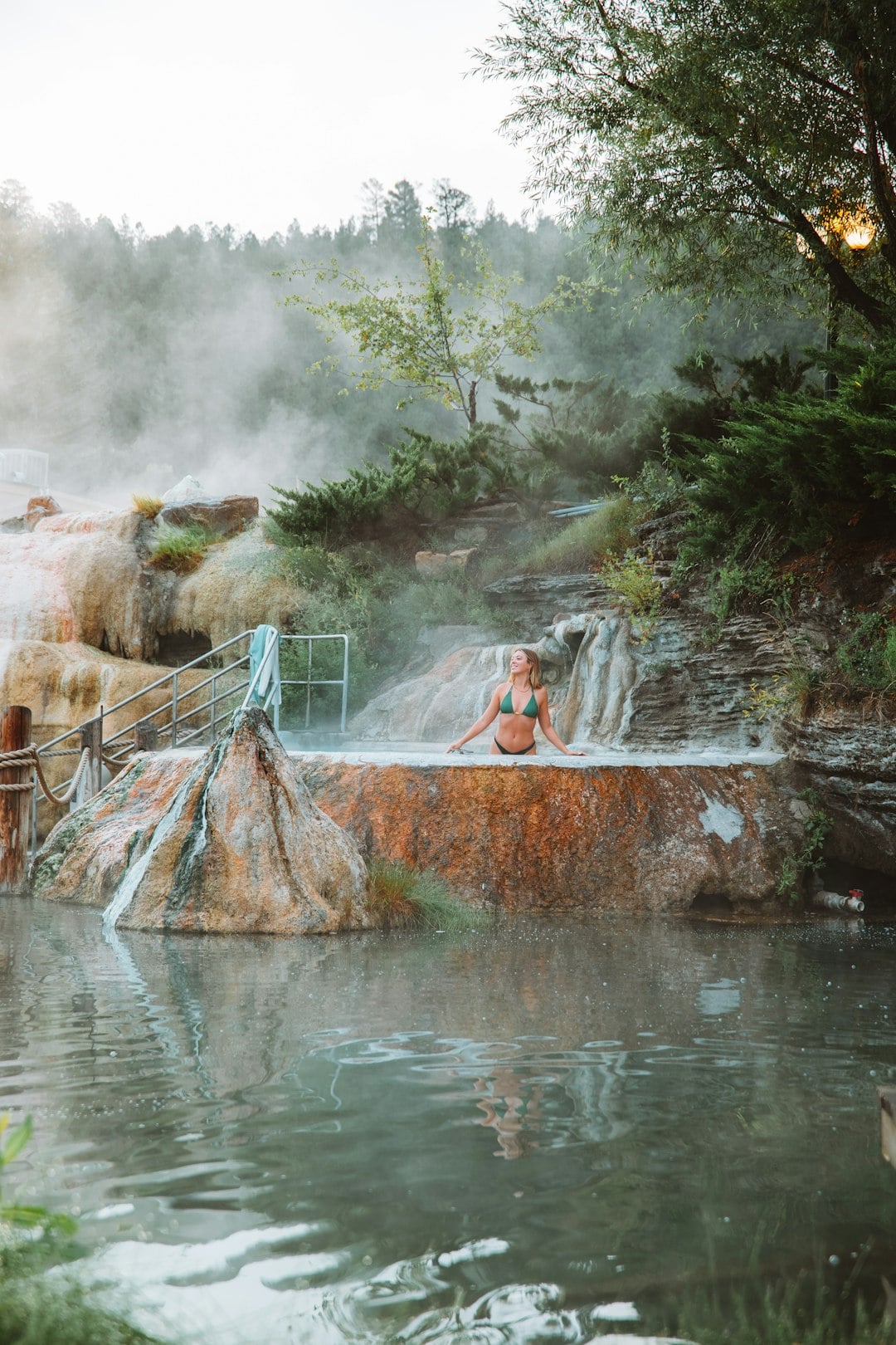 A woman in a green bikini smiles, relaxing in a steamy natural hot spring pool. It's surrounded by mossy rocks and lush greenery, with mist rising and a larger water body below.