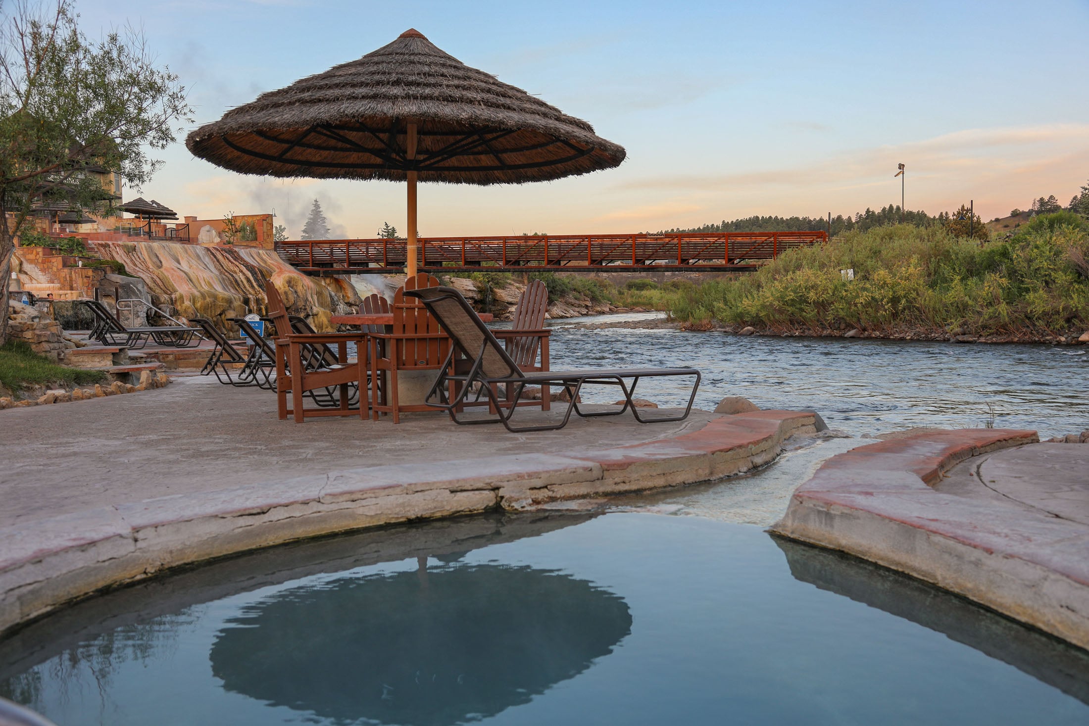 A thatched umbrella stands over lounge chairs beside hot springs pools. Steam rises from distant pools by a river and a wooden bridge, all under a sunset sky.
Transcribed text: THE SPRINGS, SULPHUR RICH.
