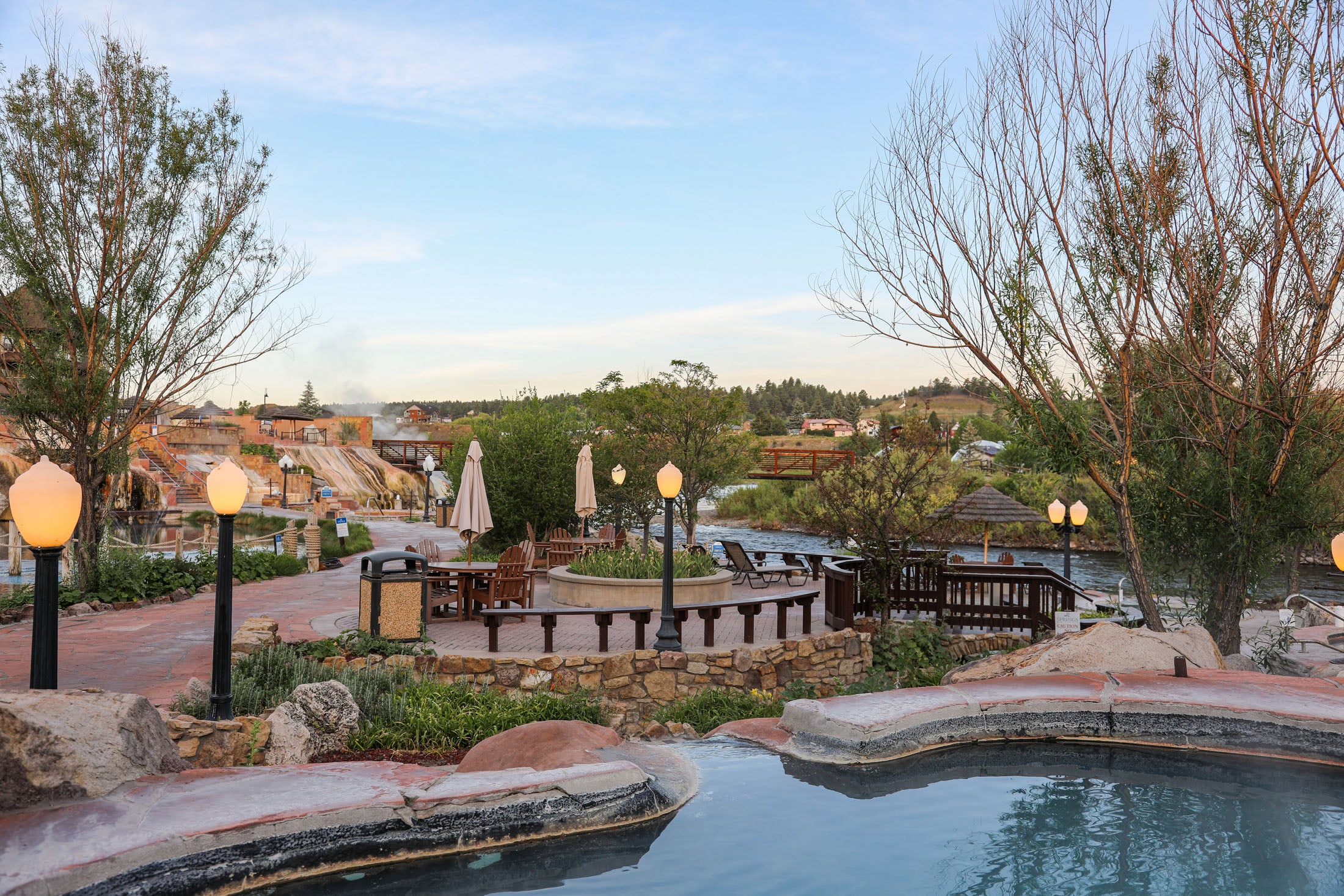 Hot springs resort pools and lit pathways lead to dining areas beside a river. Buildings and hills are visible under a pale evening sky. Text: 