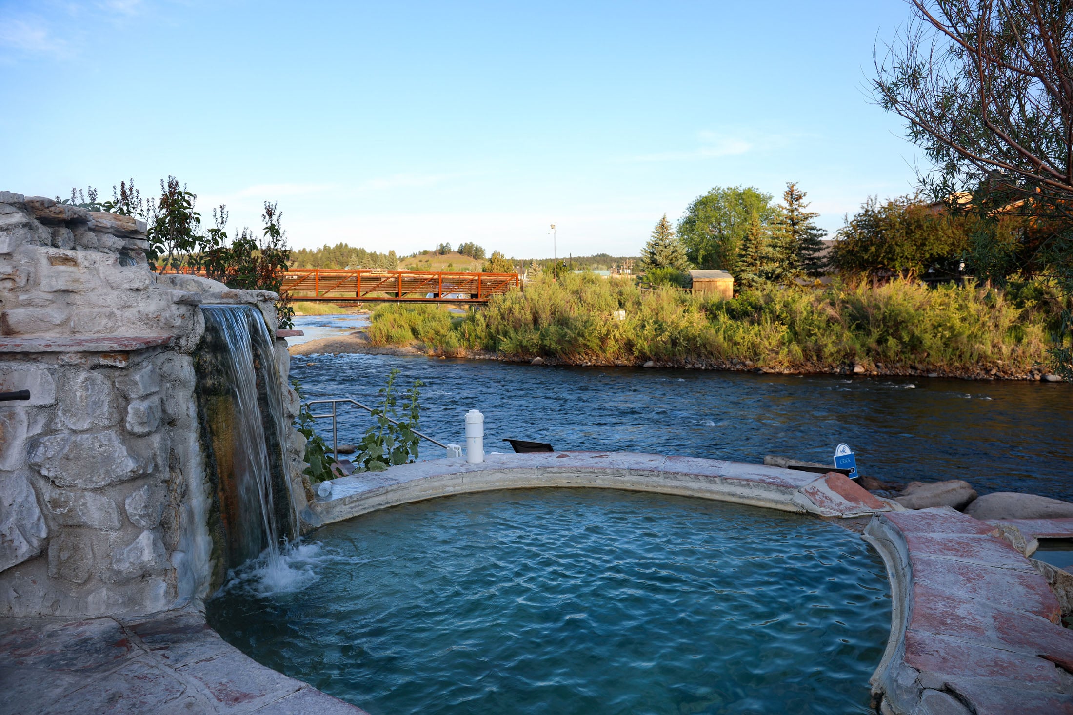 A stone hot spring pool with a waterfall sits beside a flowing river. A bridge crosses the river in the distance, surrounded by green foliage. Sign: Crick