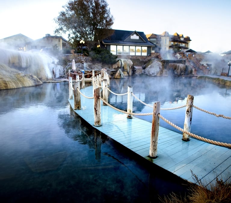 A light blue wooden boardwalk extends into a steaming natural hot spring pool, bordered by posts and rope. Buildings, steam, and mineral deposits line the background.