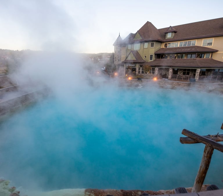 A large, turquoise-blue hot spring pool steams heavily, situated next to a multi-story hotel with stone and tan siding. A sign on the building reads: 