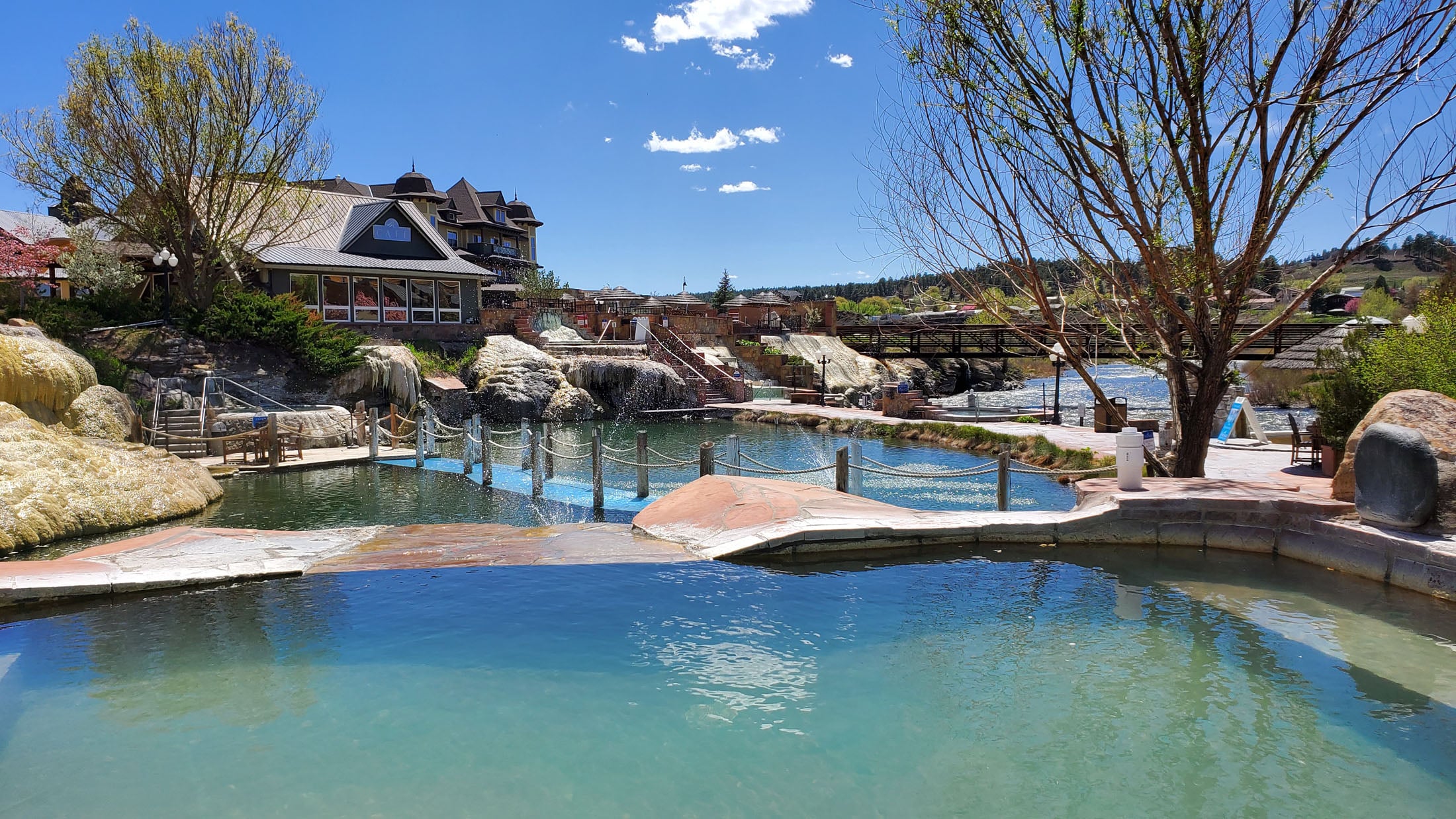 Interconnected outdoor pools and bridges flow amidst a resort building, natural rock formations, and a river under a distant bridge, all beneath a clear blue sky.