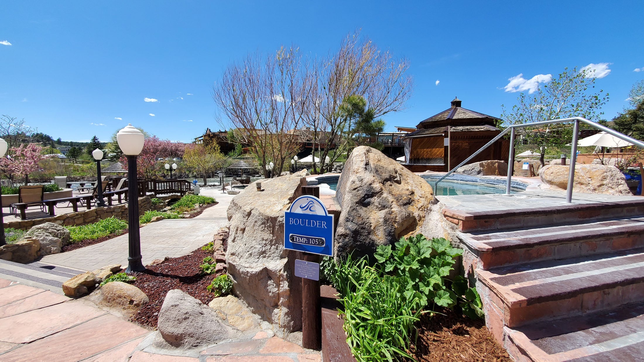 Blue sign labels a hot spring, set among boulders, stone stairs, and trees at an outdoor resort under a clear sky. Text: BOULDER TEMP: 105°F. ADULT ONLY FOOD & ENTERTAINMENT 10 AM – 11 PM DAILY (970) 586-2180.