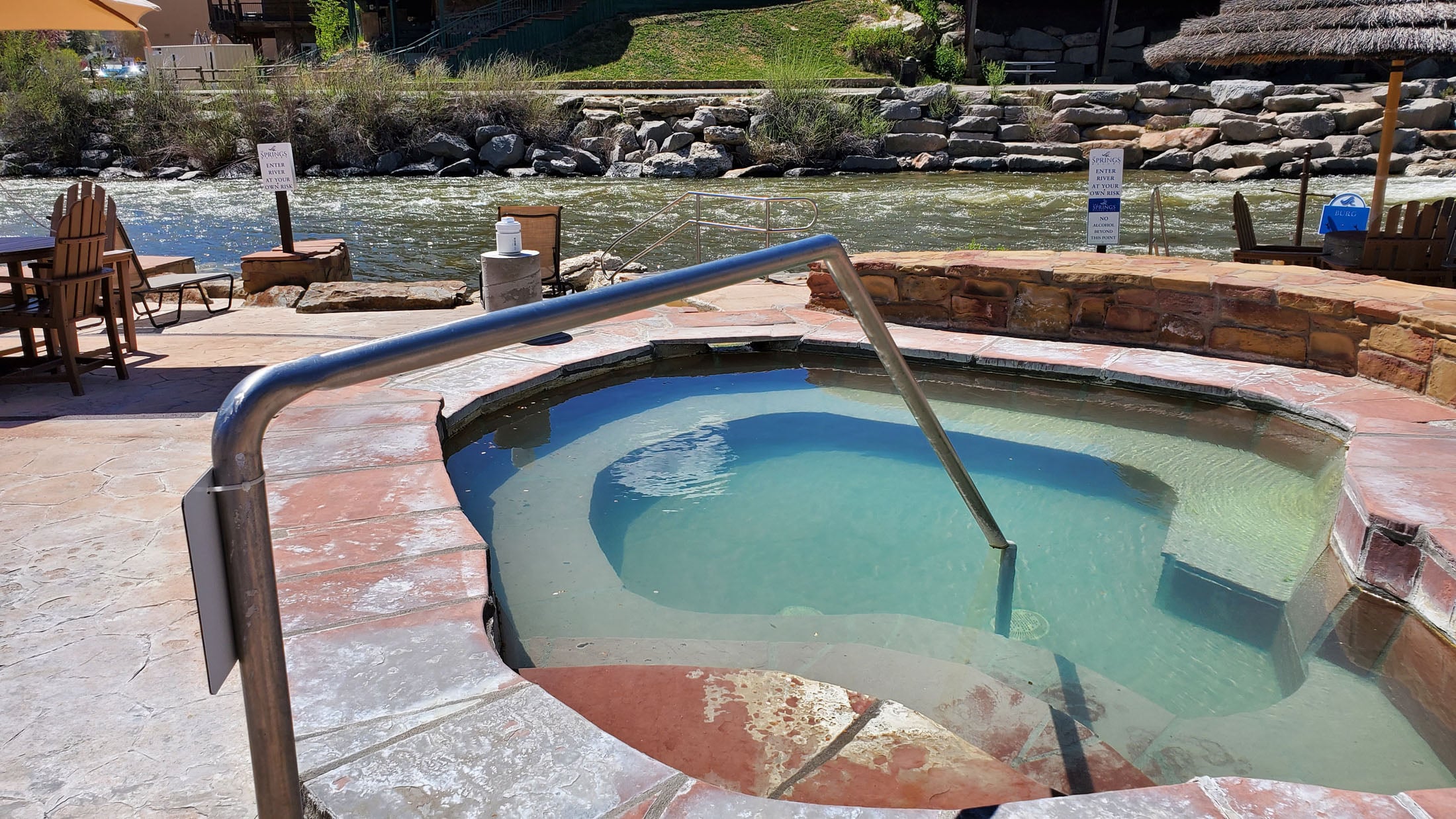 An outdoor, multi-tiered hot tub filled with clear water, featuring a metal handrail for entry, sits on a stone patio beside a flowing river. Signs read: SPRINGS ENTER RIVER AT YOUR OWN RISK NO ALCOHOL BEYOND THIS POINT.