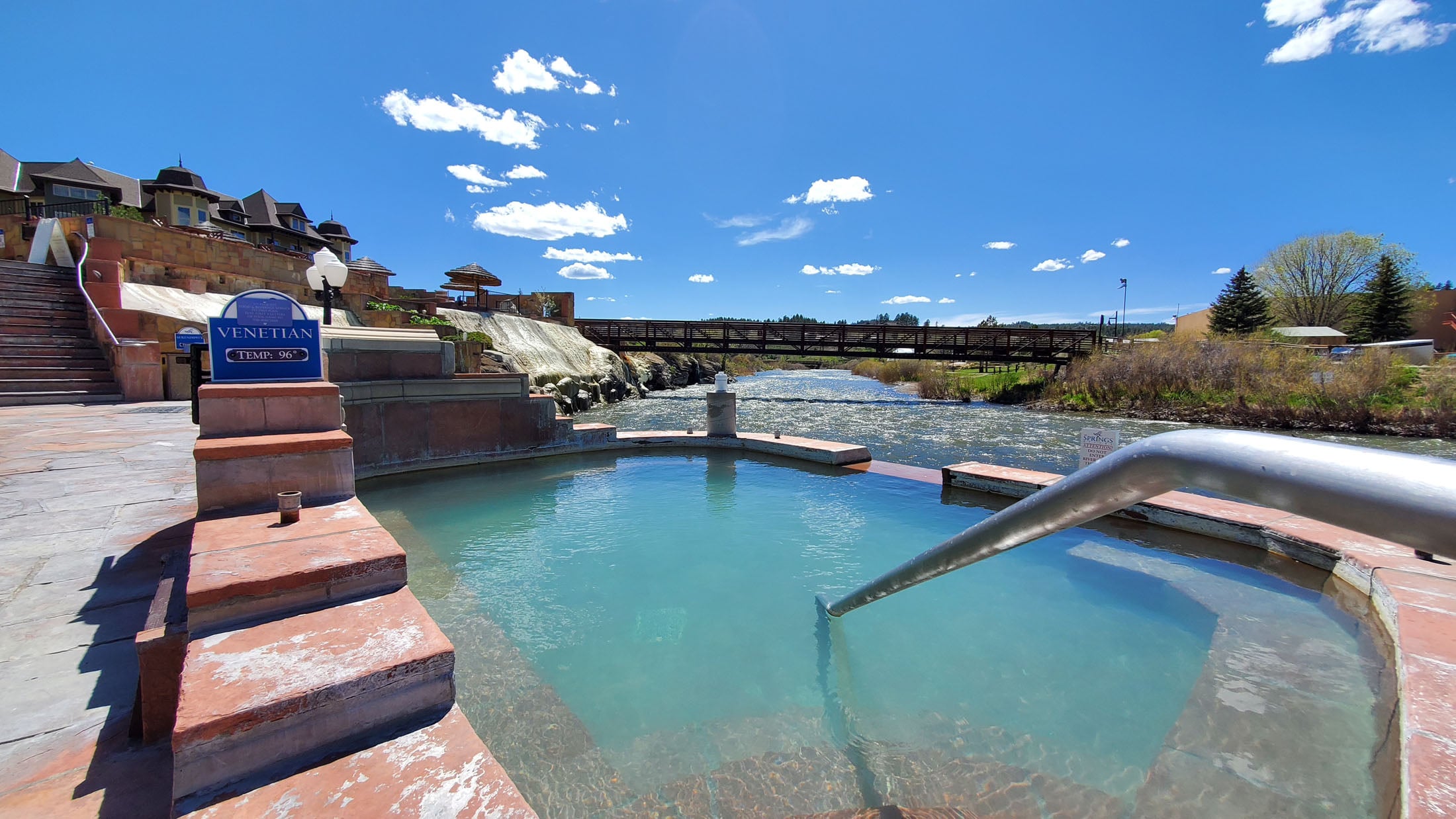 A turquoise hot spring pool with a metal handrail sits beside a flowing river, bordered by multi-tiered buildings and a wooden bridge under a bright blue sky.

Text: 
