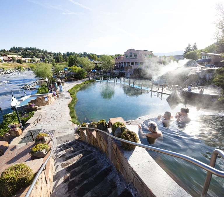 People relax in multiple steaming hot springs and a large calm pool, situated next to a fast-flowing river with distant buildings and green hills.
