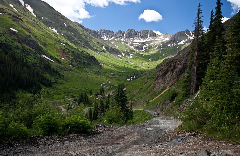Two ATVs drive down a winding dirt road through a lush green mountain valley with snow-capped peaks under a blue sky.