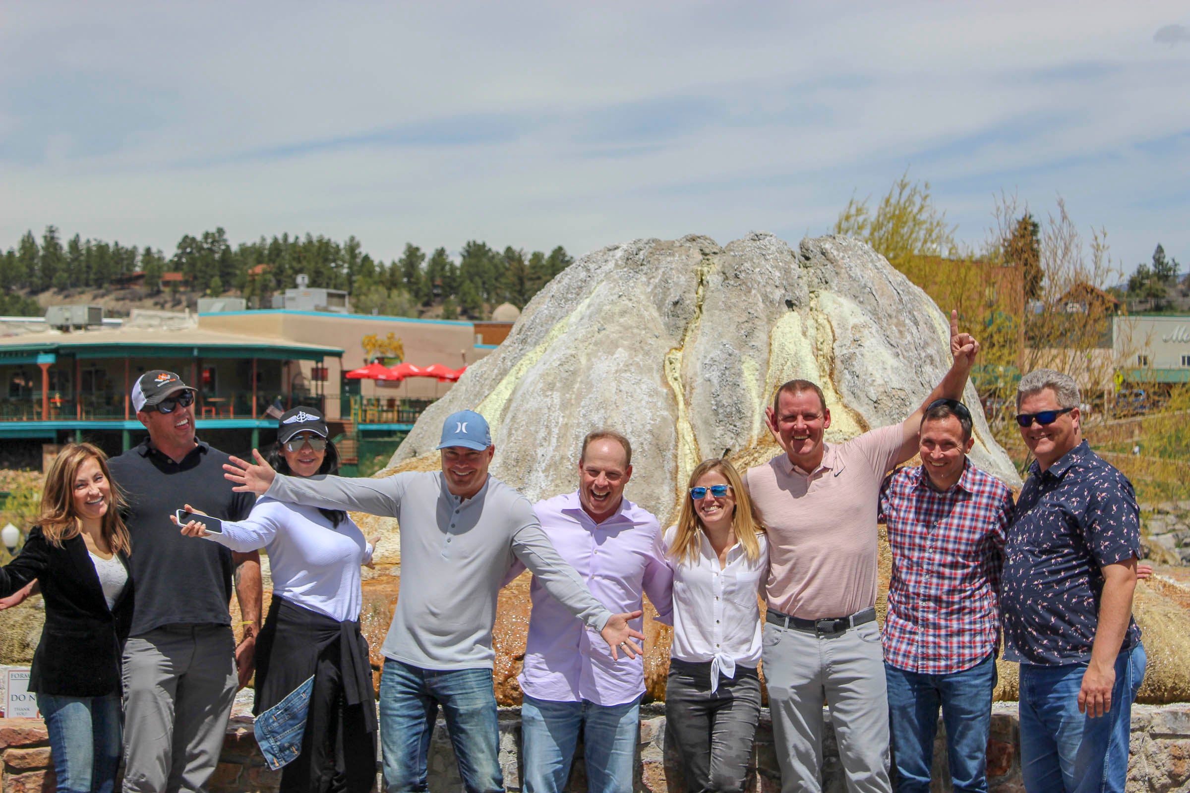 Nine adults (five men, four women) pose with outstretched arms and smiles in front of a white and yellow mineral formation. Distant buildings and pine trees are visible under a partly cloudy sky. Text: 