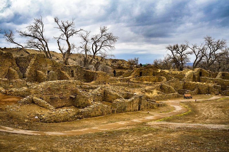 Weathered sandstone ruins stand as a complex of partially intact structures, with a dirt path winding through, surrounded by bare trees under a cloudy sky.