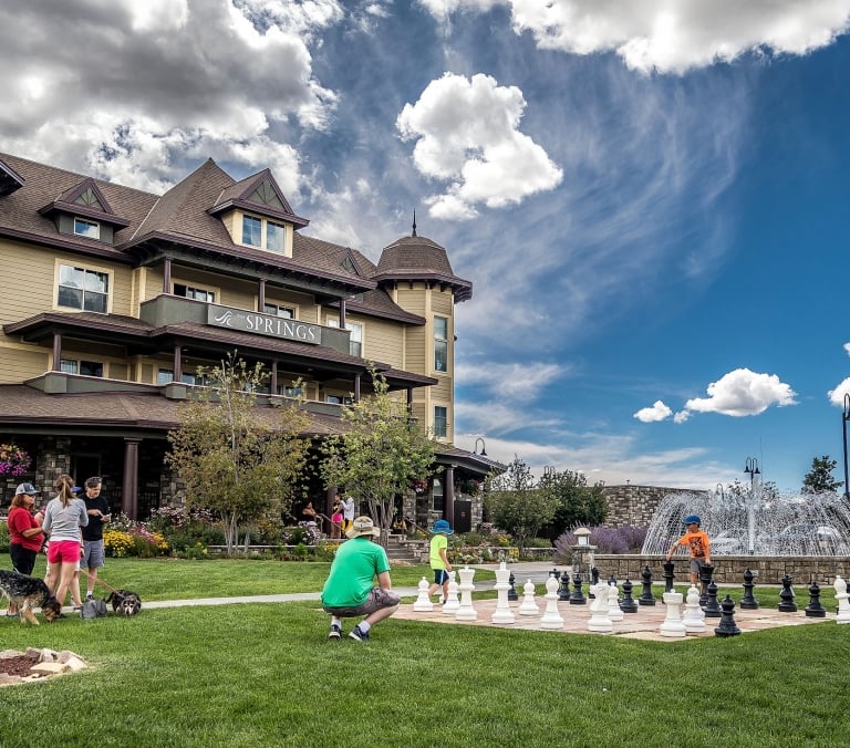 A grand hotel overlooks people playing giant chess and walking dogs on a green lawn with a fountain, under a partly cloudy sky. Text: THE SPRINGS.