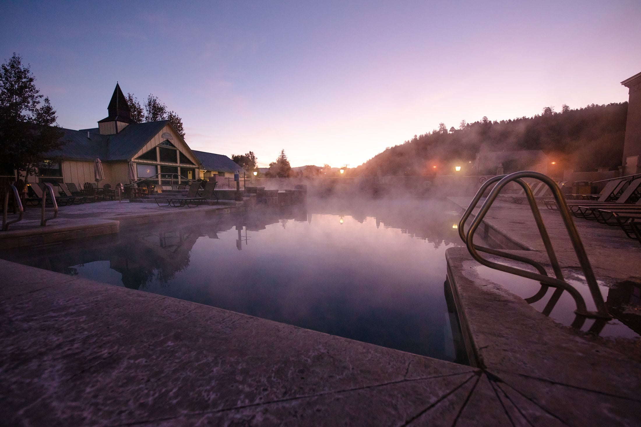 Steaming outdoor hot springs reflect a purple-pink twilight sky, flanked by a resort building and a tree-covered hill.