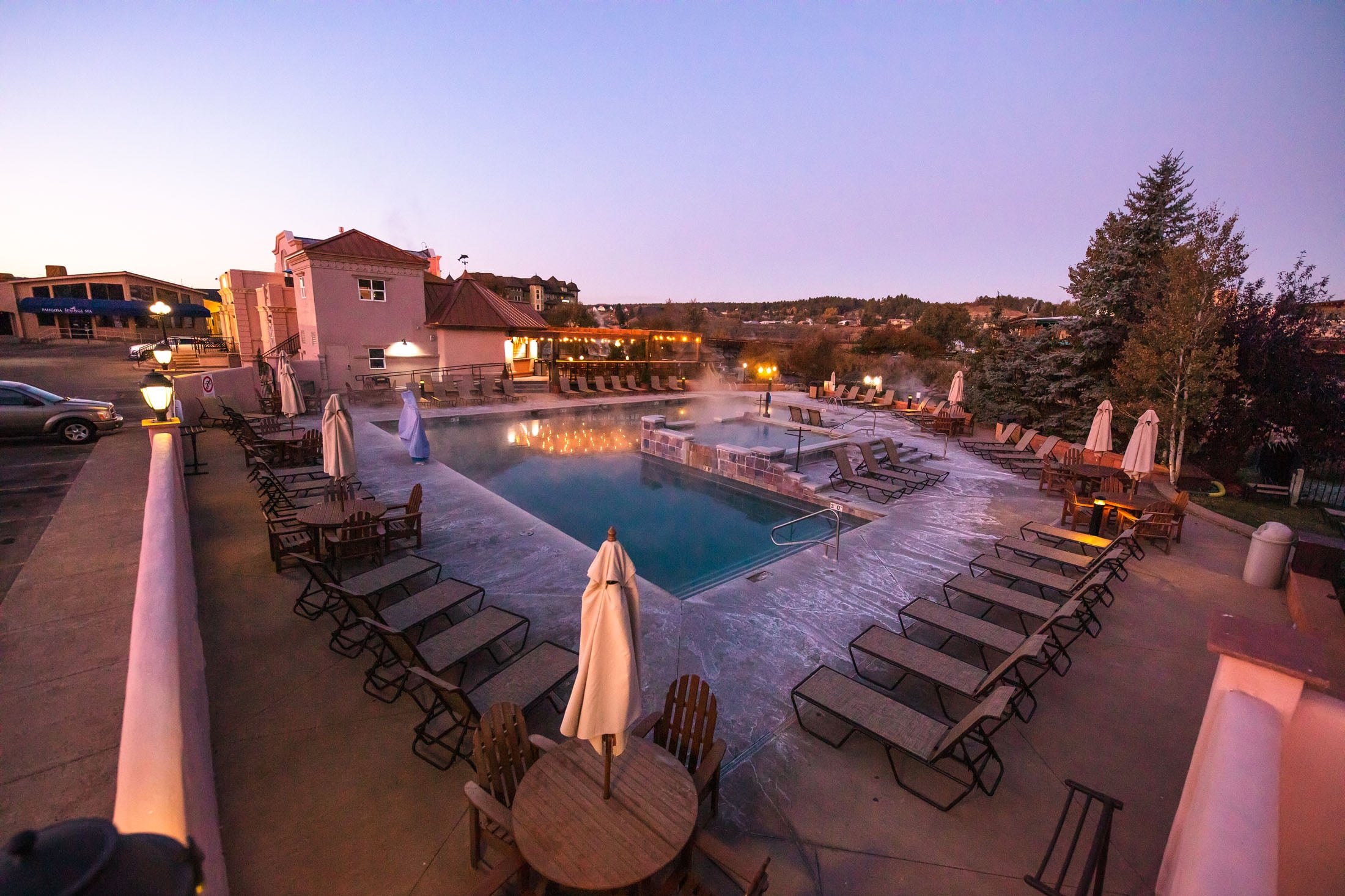 Steaming outdoor pools reflect lights, surrounded by numerous lounge chairs and closed umbrellas. Buildings, including 