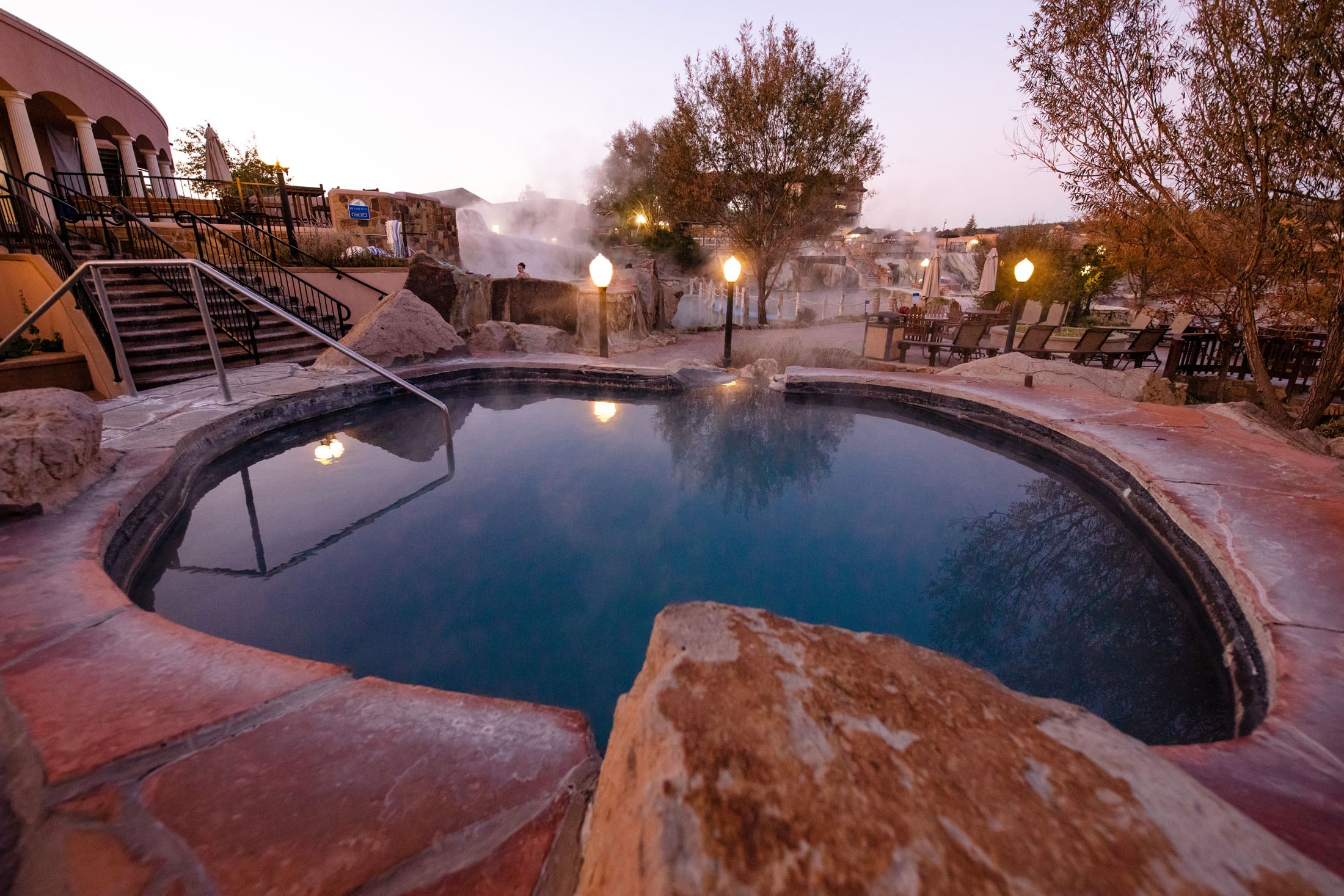A dark-blue hot spring pool reflects lamps, surrounded by flagstone, rocks, and trees. Steam rises from background pools near a building and lounge chairs under a twilight sky. Signage: NON-POTABLE NOT FOR DRINKING.