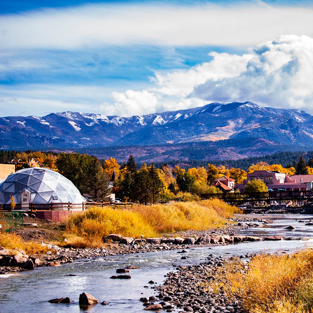 A geodesic dome building stands by a rocky river flowing through golden autumn foliage, with distant snow-capped mountains under a blue sky.