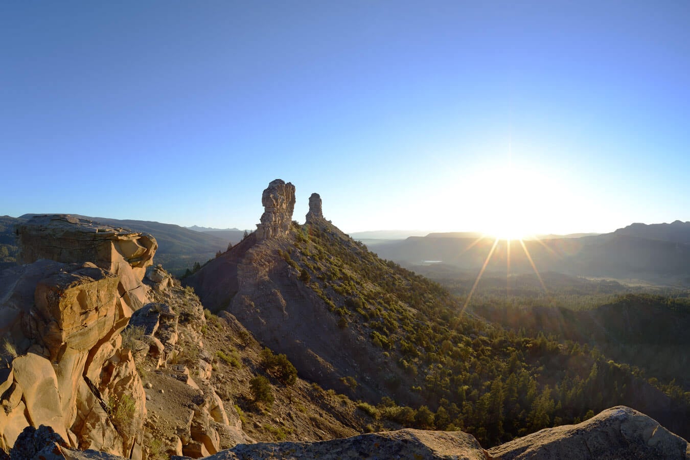 A distinctive double rock spire crowns a barren, scrub-covered hill, dominating a vast mountainous landscape as the sun creates a bright starburst on the hazy horizon.
