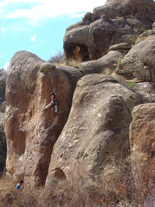 A climber ascends a large, brown rock face, tethered by a rope. Another person sits at the base amidst dry vegetation under a bright blue sky.